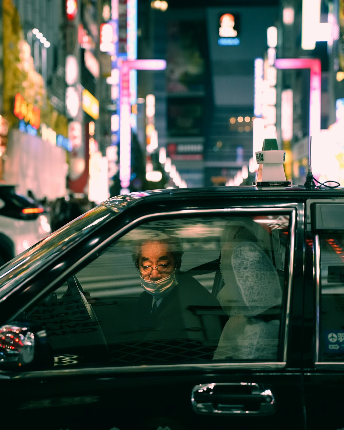 An older taxi driver navigating a city street at night.